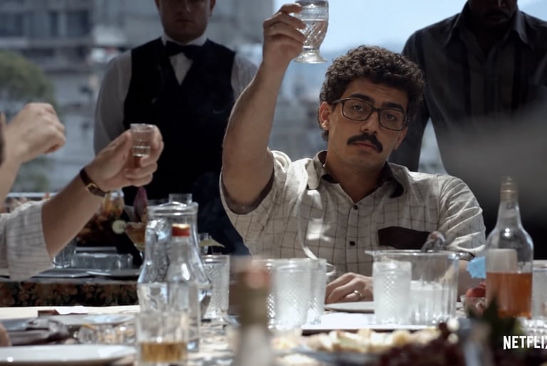 Man wearing glasses and plaid shirt raising a glass at a table with other formally dressed people at what appears to be a business meeting or formal dinner.