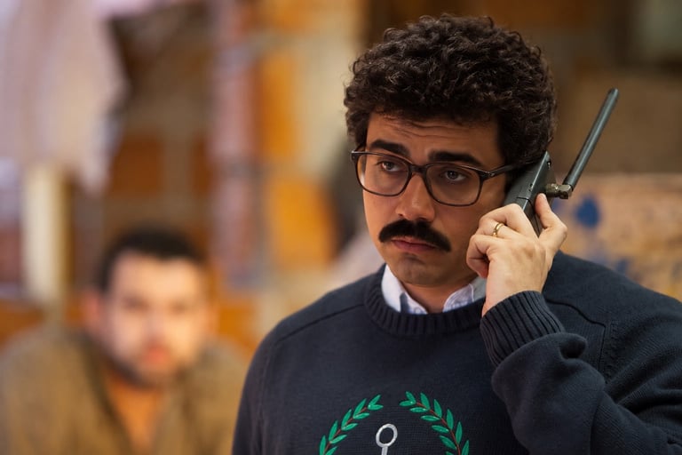 Man with mustache and glasses holding a walkie-talkie, wearing a dark sweater with a laurel wreath design