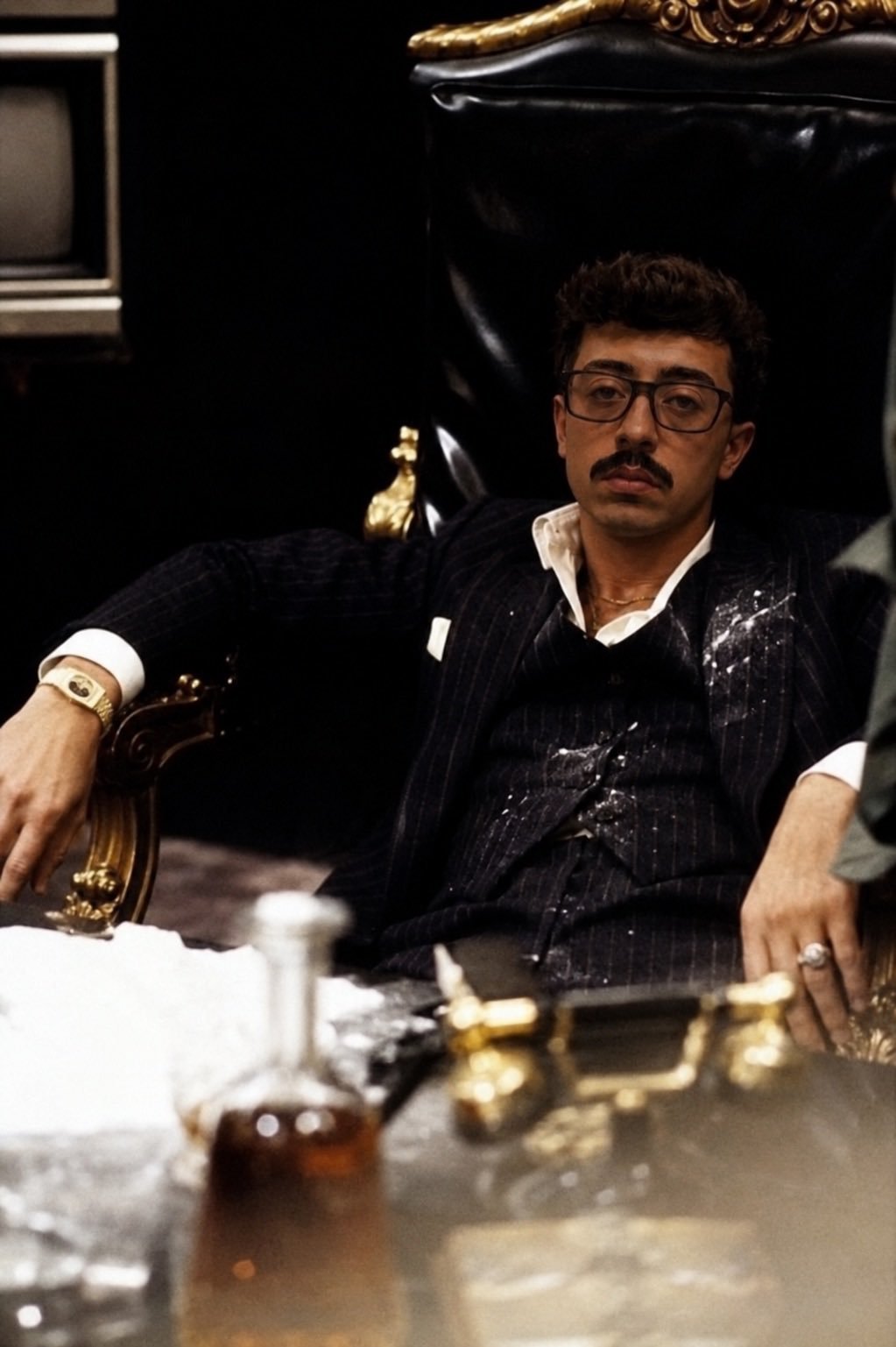 Man in pinstripe suit and glasses seated at desk with gold accessories and ornate furniture in background